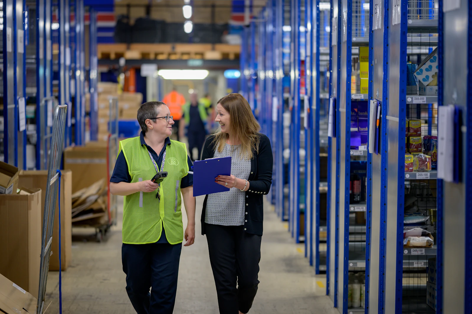 Dos trabajadoras recorren un pasillo de almacén entre estanterías con inventario; una lleva un escáner y la otra una carpeta.