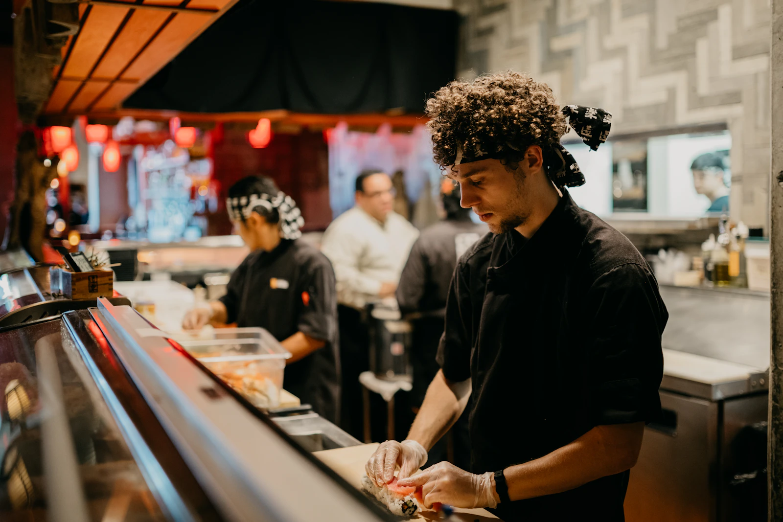 Cocinero preparando un platillo en la barra de un restaurante mientras el equipo trabaja al fondo.